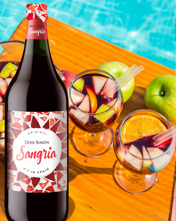 A bottle of Don Simón Sangria sits on a wooden table beside two glasses filled with sangria, sliced fruit, and straws. Green apples and a swimming pool are in the background, suggesting a refreshing summer scene.