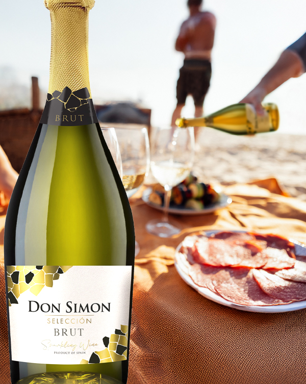 A bottle of Don Simon Selección Brut sparkling wine stands on an orange picnic blanket with plates of sliced meat and glasses, as a person pours wine at a beach picnic scene in the background.